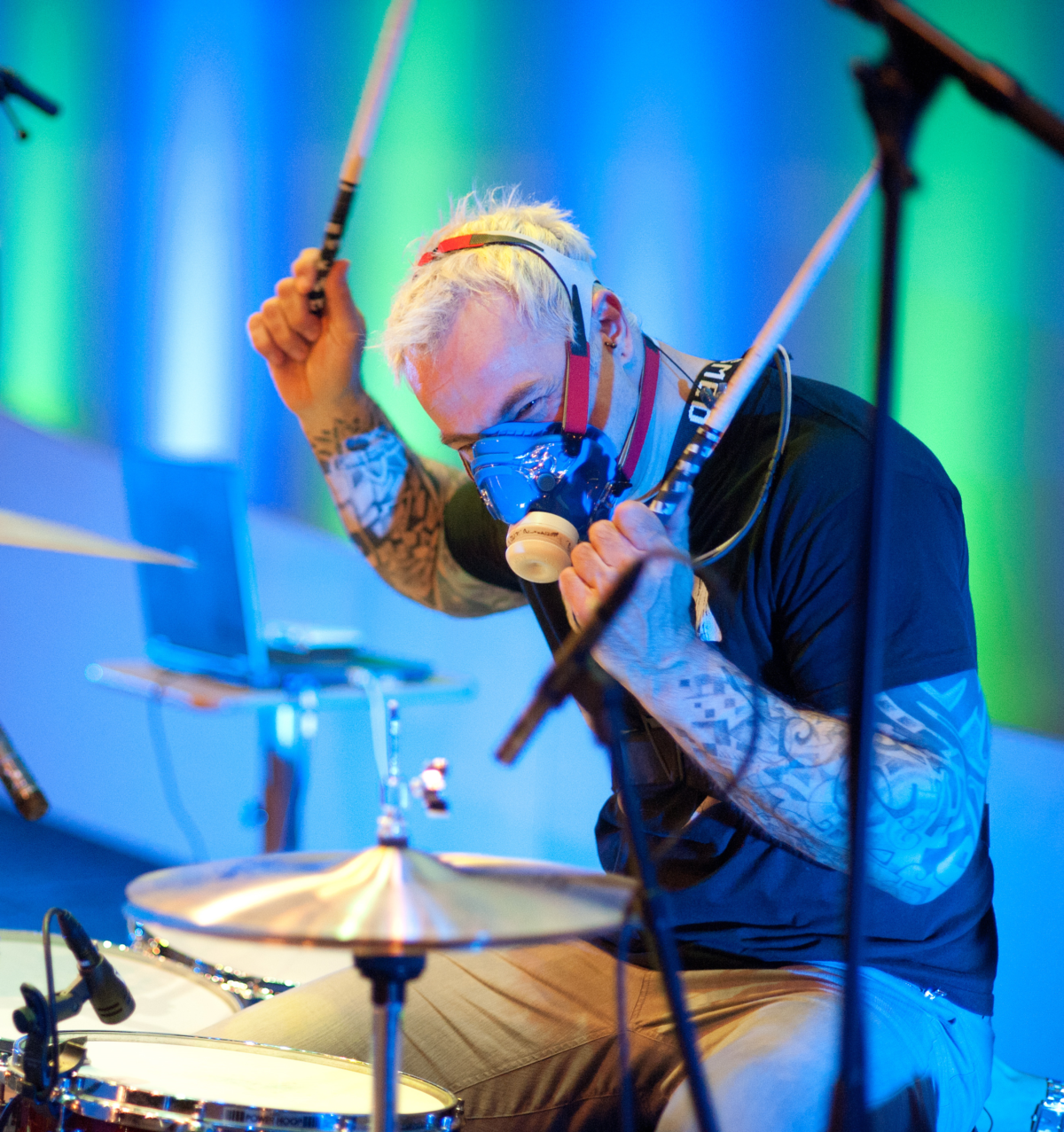 A man with short blonde hair plays drums wearing an oxygen mask